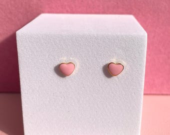 Pode incluir: Um par de brincos de pressão em forma de coração rosa com detalhes dourados. Os brincos são exibidos em uma plataforma branca.