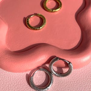 Può includere: Due paia di orecchini a cerchio, un paio è dorato e l'altro è argentato. Entrambe le paia hanno un design testurizzato.