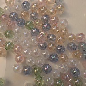 May include: A collection of round, spherical beads with an iridescent, pearlescent finish. The beads are scattered and feature a variety of soft pastel colors, including light blue, pale pink, mint green, and translucent white, each with a central hole.