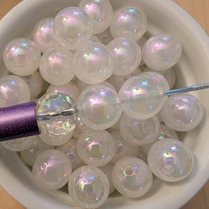 May include: A collection of round, white beads with an iridescent finish, featuring a pearlescent sheen that reflects subtle rainbow colors. One bead is faceted and attached to a purple metal tool, while others are loose in a white bowl.