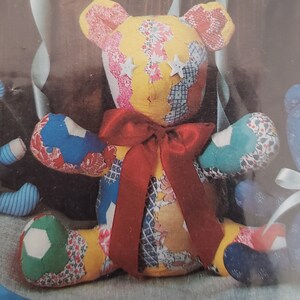 May include: A patchwork teddy bear with a red bow. The bear is made of different fabrics in various colors and patterns, including yellow, blue, green, and pink. The bear has white button eyes and a stitched smile.