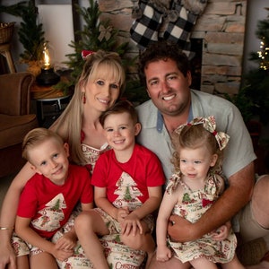 May include: A family of four wearing matching Christmas-themed outfits. The parents are sitting on the floor with their two children. The children are wearing red shirts with a Christmas tree design and the parents are wearing cream-colored shirts with a Christmas tree design. The family is smiling and looking at the camera.