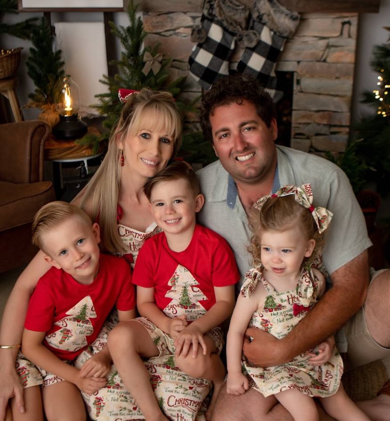 May include: A family of four wearing matching Christmas-themed outfits. The parents are sitting on the floor with their two children. The children are wearing red shirts with a Christmas tree design and the parents are wearing cream-colored shirts with a Christmas tree design. The family is smiling and looking at the camera.