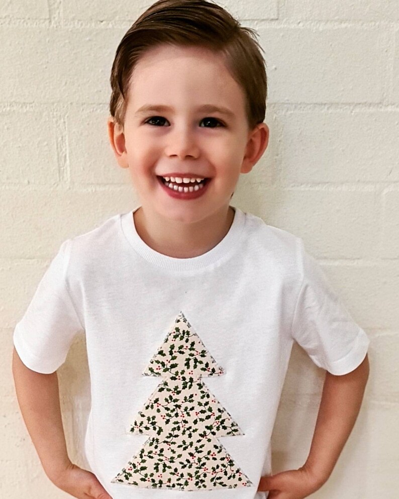 May include: A white t-shirt with a festive Christmas tree design. The tree is made of fabric with a green and red holly pattern.