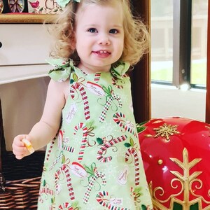 May include: A young girl wearing a green dress with a candy cane pattern. The dress has red and white candy canes, holly leaves, and the words "Happy Holidays" on it. The girl is smiling and has blonde curly hair. She is wearing a red and white bow in her hair.