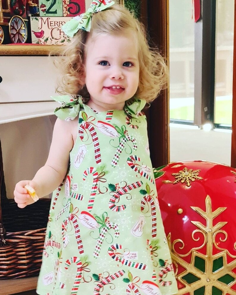 May include: A young girl wearing a green dress with a candy cane pattern. The dress has red and white candy canes, holly leaves, and the words "Happy Holidays" on it. The girl is smiling and has blonde curly hair. She is wearing a red and white bow in her hair.
