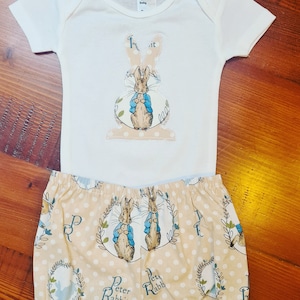 May include: A white onesie with a Peter Rabbit appliqué on the front. The onesie is paired with beige trousers with a Peter Rabbit print.