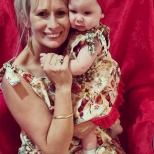 May include: A woman and a young girl wearing matching Christmas-themed dresses. The woman is holding the girl in her arms. The girl is wearing a red bow in her hair. The background is a red wall.