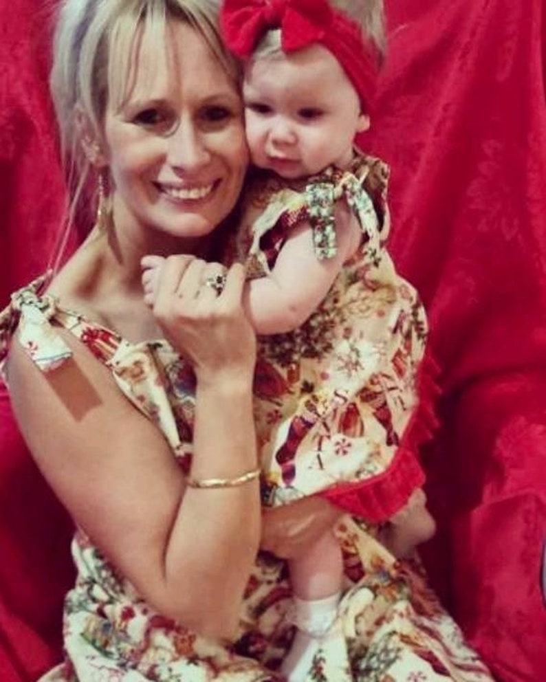 May include: A woman and a young girl wearing matching Christmas-themed dresses. The woman is holding the girl in her arms. The girl is wearing a red bow in her hair. The background is a red wall.