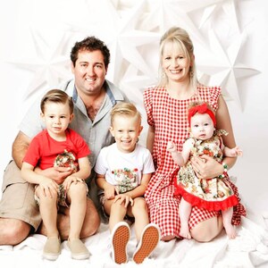 May include: A family of four poses for a Christmas photo. The parents are wearing casual clothing, while the two children are dressed in festive outfits. The background is decorated with white stars.