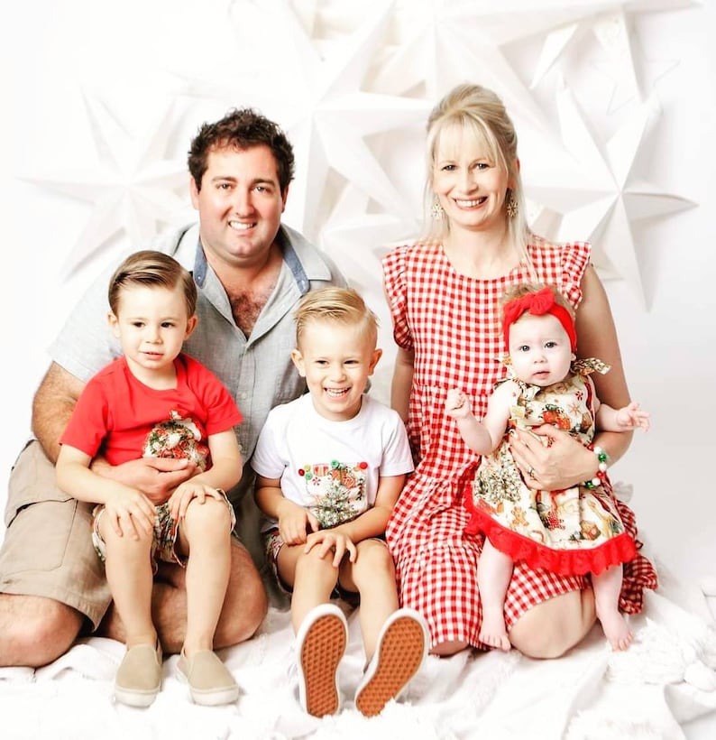 May include: A family of four poses for a Christmas photo. The parents are wearing casual clothing, while the two children are dressed in festive outfits. The background is decorated with white stars.