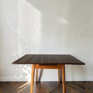 May include: A rectangular wooden table featuring a dark brown, vertically striped tabletop surface and four light brown, tapered wooden legs. The table has a flat, smooth top with a distinct grain pattern and a sturdy, simple frame construction.