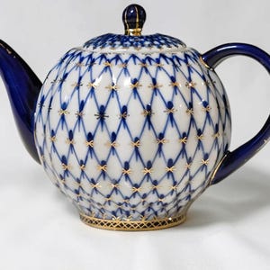 Könnte beinhalten: Eine runde Teekanne aus Porzellan mit weißem Grund, blauem Gittermuster und goldenen Akzenten. Die Kanne hat einen dunkelblauen Ausguss, einen passenden Henkel und einen Deckel mit kleinem Knauf. Der Boden ist mit einer goldenen Bordüre verziert.