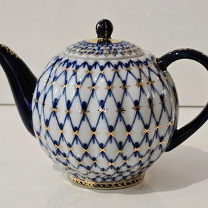 Könnte beinhalten: Eine weiße Porzellan-Teekanne mit blauem und goldenem Gittermuster. Die Teekanne hat eine runde Form, einen gebogenen Ausguss und einen dunkelblauen Griff und Deckel. Die goldenen Akzente verleihen diesem Dekorationsstück Eleganz.