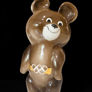 Peut inclure: Une figurine d'ours en céramique marron brillant avec un museau blanc, un nez noir et une expression souriante. L'ours porte une ceinture jaune ornée du symbole des anneaux olympiques blancs. La figurine a des oreilles rondes et une finition lisse.