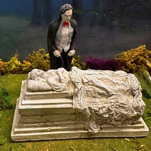 May include: A miniature scene featuring a figure in a black cape and red bow tie, standing over a stone sarcophagus. The sarcophagus has a sculpted figure draped in cloth. The background shows a misty forest and green moss.