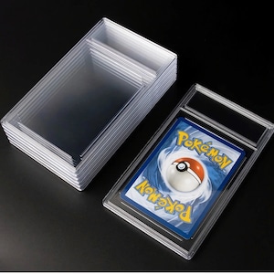 May include: Clear plastic card holders and a graded Pokémon card. The graded card is encased in a rigid, transparent holder. A stack of similar, empty holders is also visible, suggesting a collection or storage solution for trading cards.