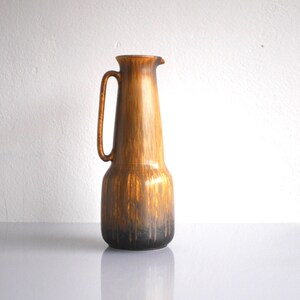 May include: A tall, brown ceramic pitcher with a handle and a spout. The pitcher has a textured surface with a gradient of colors, transitioning from light brown at the top to dark brown at the base. The pitcher is likely used for serving beverages.