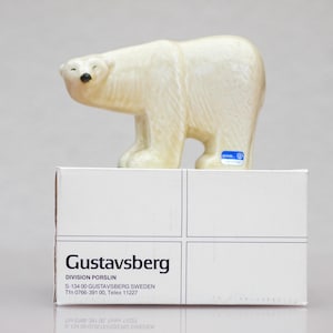 May include: A cream-colored ceramic polar bear figurine with a glossy finish and a small blue sticker on its side, positioned atop a white rectangular cardboard box labeled with the text Gustavsberg Division Porslin S-134 00 Gustavsberg Sweden.