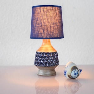 Puede incluir: Una lámpara de mesa vintage con una pantalla azul marino y una base de cerámica decorada con motivos florales azules. Una pequeña figura de pájaro de cerámica con detalles azules y marrones está junto a la lámpara.