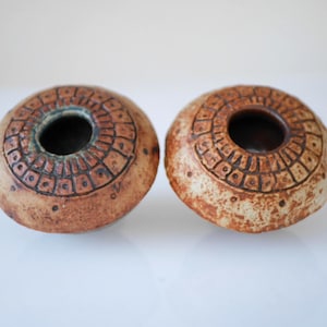 Puede incluir: Dos candeleros redondos de cerámica hechos a mano. Los candeleros son de color marrón cálido con un diseño texturizado y estampado alrededor de la parte superior. Cada uno tiene una abertura oscura y circular en el centro.