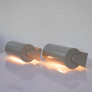 May include: Two cylindrical, taupe-coloured lamps with a rectangular shade attached to the base. The lamps are lit, casting a warm glow. The lamps are on a white surface, with a white wall in the background. The lamps are a modern design.