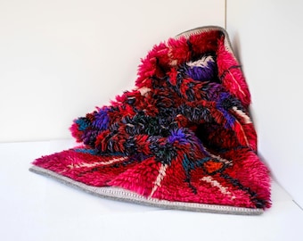 May include: A thick, shaggy rug featuring a vibrant color palette of deep magenta, dark purple, black, and hints of white. The rug has a high-pile texture with a folded corner, revealing a gray fabric backing along the finished, stitched edges.