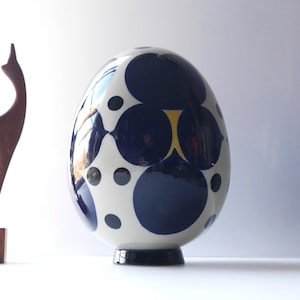 Puede incluir: Un jarrón de cerámica blanco con forma de huevo con un patrón geométrico azul y blanco. El jarrón tiene una base redonda y está decorado con un patrón repetitivo de círculos y óvalos.