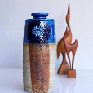 以下が含まれることがあります： 青い上部と、質感のある茶色とベージュのボディを持つ、背の高いセラミック花瓶。青い部分には、円形のスターバーストデザインがあります。鳥と鹿の木彫りの彫刻が背景にあります。