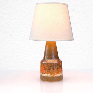 Puede incluir: Lámpara de mesa de cerámica con pantalla blanca. La base de la lámpara es marrón texturizado con detalles en naranja. La lámpara es una lámpara decorativa.