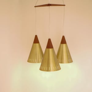Peut inclure: Un luminaire suspendu de style moderne du milieu du siècle avec trois abat-jours en forme de cône. Les abat-jours sont dorés avec un dessus en bois marron et un design à fentes. Le luminaire est suspendu à un carré en bois.