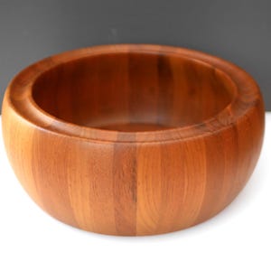 Puede incluir: Un cuenco redondo de madera de color marrón rojizo cálido. El cuenco tiene un diseño amplio y abierto con un acabado liso y pulido. La veta de la madera es visible, lo que se suma a su estética natural. Este cuenco es adecuado para servir ensaladas o como pieza decorativa.