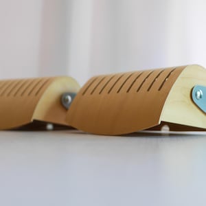 May include: Two wooden shoe trees in a light brown colour. Each tree features a curved top with evenly spaced slits and a light blue metal handle. The shoe trees are designed to maintain the shape of shoes.