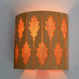 Puede incluir: Aplique de pared de madera con un patrón de hojas recortadas, que emite un cálido resplandor anaranjado. El aplique es de madera marrón claro y está montado en una pared blanca. Es una lámpara decorativa.