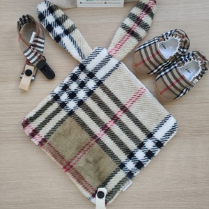 Op de afbeelding: Een beige en bruin geruite babydeken met een bijpassend paar babyschoentjes. De schoentjes zijn gemarkeerd met "3-6m". De deken heeft een zwarte knoop en een bijpassende clip.