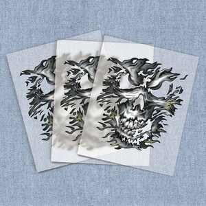 May include: Three sheets of white transfer paper with a black and white skull design printed on them. The skull is surrounded by flames.