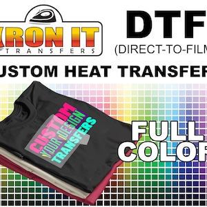 May include: A black t-shirt with a colorful design that says "Custom Your Design Transfers" on it. The t-shirt is stacked on top of two other t-shirts, one beige and one red. The image is set against a background of a color chart with a variety of colors.