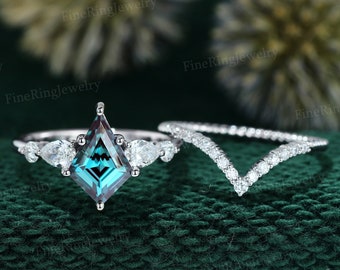 May include: A two-piece engagement ring set. The primary ring features a kite-shaped, teal-colored gemstone, flanked by pear-shaped stones. A curved band, set with small, clear stones, accompanies the main ring. The rings are set against a dark green background.