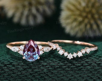 May include: A pear-shaped, iridescent gemstone ring set in rose gold with a matching band. The ring features a halo of smaller diamonds around the center stone and the band has a delicate, curved design with small diamonds.