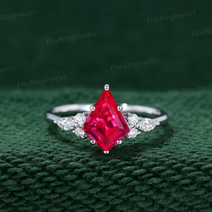 May include: A silver ring with a striking, kite-cut, red gemstone at its centre. The ring is embellished with small, clear gemstones. The background is a dark green, textured fabric. The ring is a piece of jewellery.