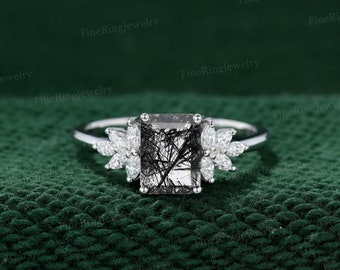 May include: A silver ring with a square-shaped black and clear gemstone surrounded by smaller clear gemstones. The ring is on a green fabric background.
