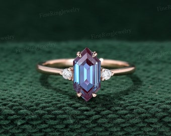 May include: A rose gold engagement ring with a hexagonal alexandrite gemstone and two smaller diamond accents. The ring is set on a green fabric background.