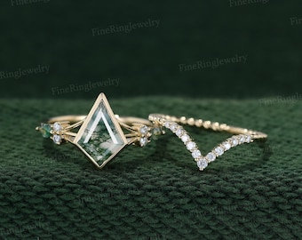 May include: Two gold rings on a green background. One ring features a large, geometric, green stone with a unique pattern. The second ring is a V-shaped band with small, clear stones. The rings are set against a dark green, textured backdrop.