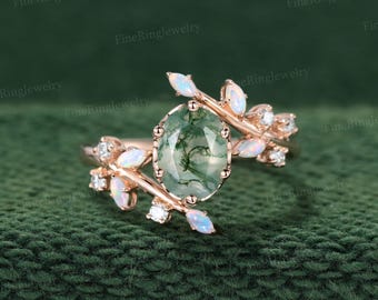 May include: A rose gold ring featuring a central oval-cut moss agate stone with green inclusions. The band is designed as a branch with small marquise-cut opals and tiny round white diamonds set along the metal leaves and stems.