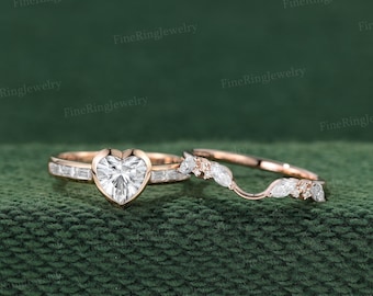 May include: A rose gold ring set featuring a heart-shaped center stone flanked by baguette-cut side stones, paired with a curved band adorned with marquise-cut and round stones. The metal has a warm, metallic finish throughout the design.