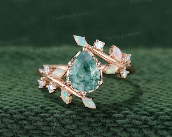 May include: A rose gold ring featuring a central pear-shaped moss agate stone, accented with marquise-cut opals and small round white stones arranged in a branch-like design along the band, creating a botanical-inspired jewelry piece.
