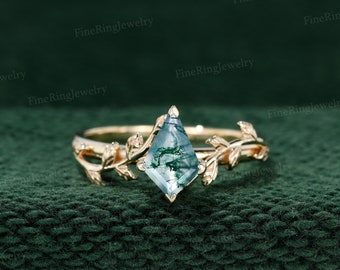 Puede incluir: Un anillo de oro con una gema de ágata musgosa en forma de cometa, con inclusiones verdes y blancas. El anillo tiene una banda decorada con motivos de hojas y vides. El fondo es una tela verde oscuro texturizada.
