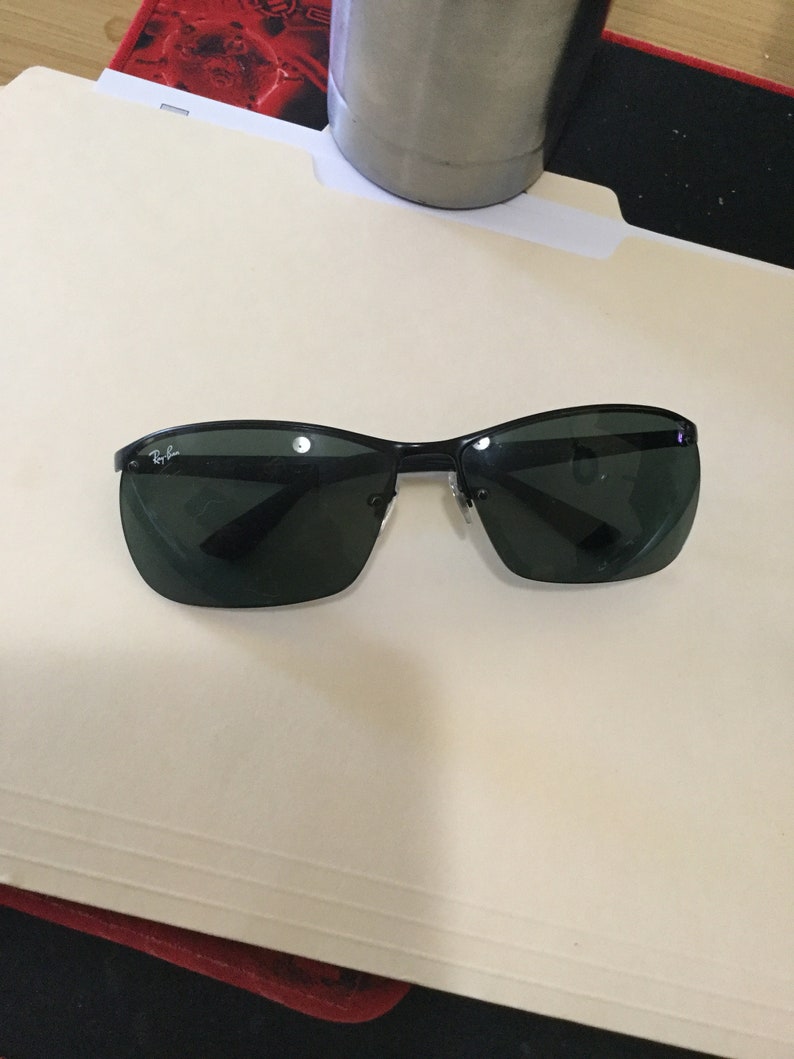 K&ouml;nnte beinhalten: Schwarze Ray-Ban Sonnenbrille mit dunkelgr&uuml;nen Gl&auml;sern. Die Sonnenbrille hat einen d&uuml;nnen, schwarzen Rahmen.