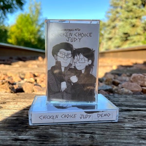 May include: A cassette tape with a clear plastic case and a black and white illustration of two people on the front. The text "Introducing Chicken Choke Judy" is printed on the front of the tape. The text "Chicken Choke Judy Demo" is printed on the bottom of the tape.