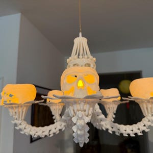 May include: A white chandelier featuring a central skull and four additional skulls on each arm. The structure is composed of interconnected vertebrae-like segments, with each skull illuminated from within by a warm, glowing light source.